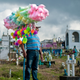 A man holds a stick with bags of cotton candy and fuzzy toys balanced on it. He's standing in a cemetery.